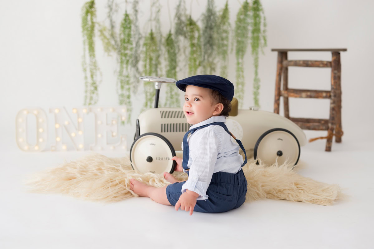 Capture the charm of your 9 to 10-month to 1-year-old with Anega Bawa's sitter photography in Delhi & Gurgaon. Your little one sits on the floor wearing a cute cap beside a toy scooter, with a leafy fringe and name frame on the floor, set against a serene white backdrop.