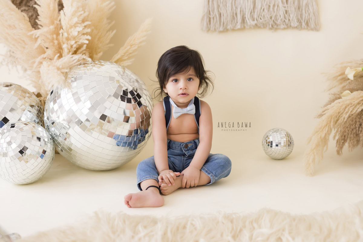 Seated amidst twinkling disco balls and surrounded by rustic brown shrubs against a serene beige backdrop, the 10-month to 1-year-old boy dons a charming blue bow. This delightful scene, captured during his baby pre-birthday photoshoot by Anega Bawa in Delhi, Gurgaon, and Noida, exudes a playful yet serene ambiance, celebrating the joyous spirit of infancy.