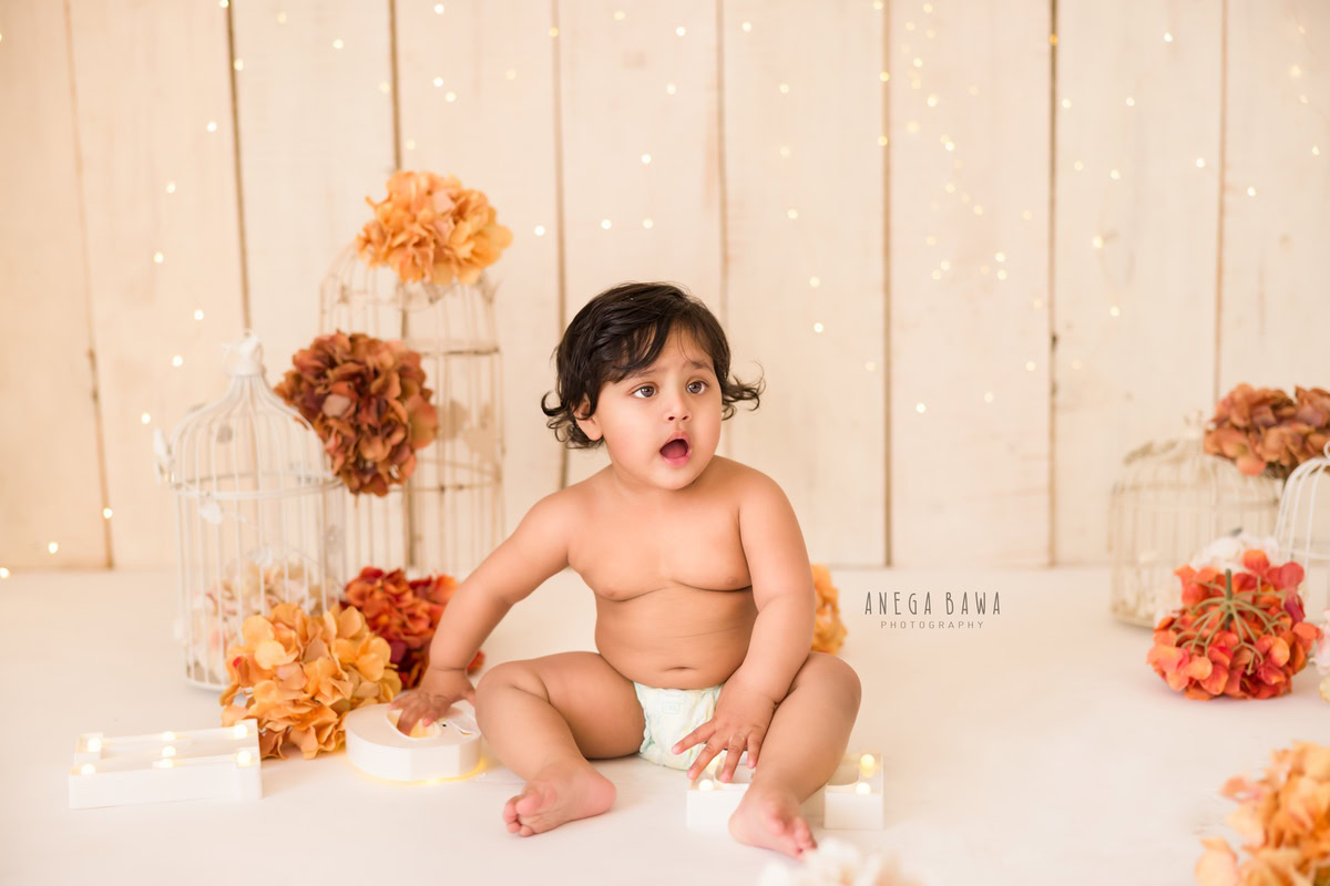 In the enchanting glow of fairy lights, surrounded by whimsical white castles and adorned with vibrant orange and brown flowers, the adorable 9-month to 1-year-old boy sits gracefully on the floor. Against a soothing beige backdrop, this captivating scene captures the essence of innocence and wonder. This charming moment was immortalized during the baby pre-birthday photoshoot by Anega Bawa, spanning across Delhi, Gurgaon, and Noida.