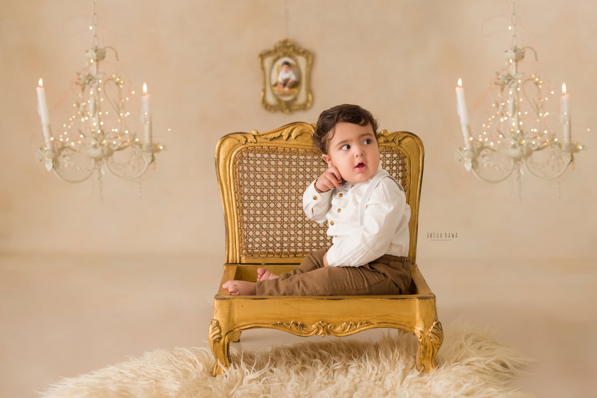 In this enchanting toddler photoshoot by Anega Bawa in Delhi, Gurgaon, and Noida, a 9 to 10-month-old boy sits gracefully on a golden wooden chair. The beige backdrop provides a subtle canvas for the scene, while a candle stand and a photo frame on the wall add decorative accents. With his innocent charm and curious expression, the boy captures the essence of childhood, creating a timeless moment to cherish.
