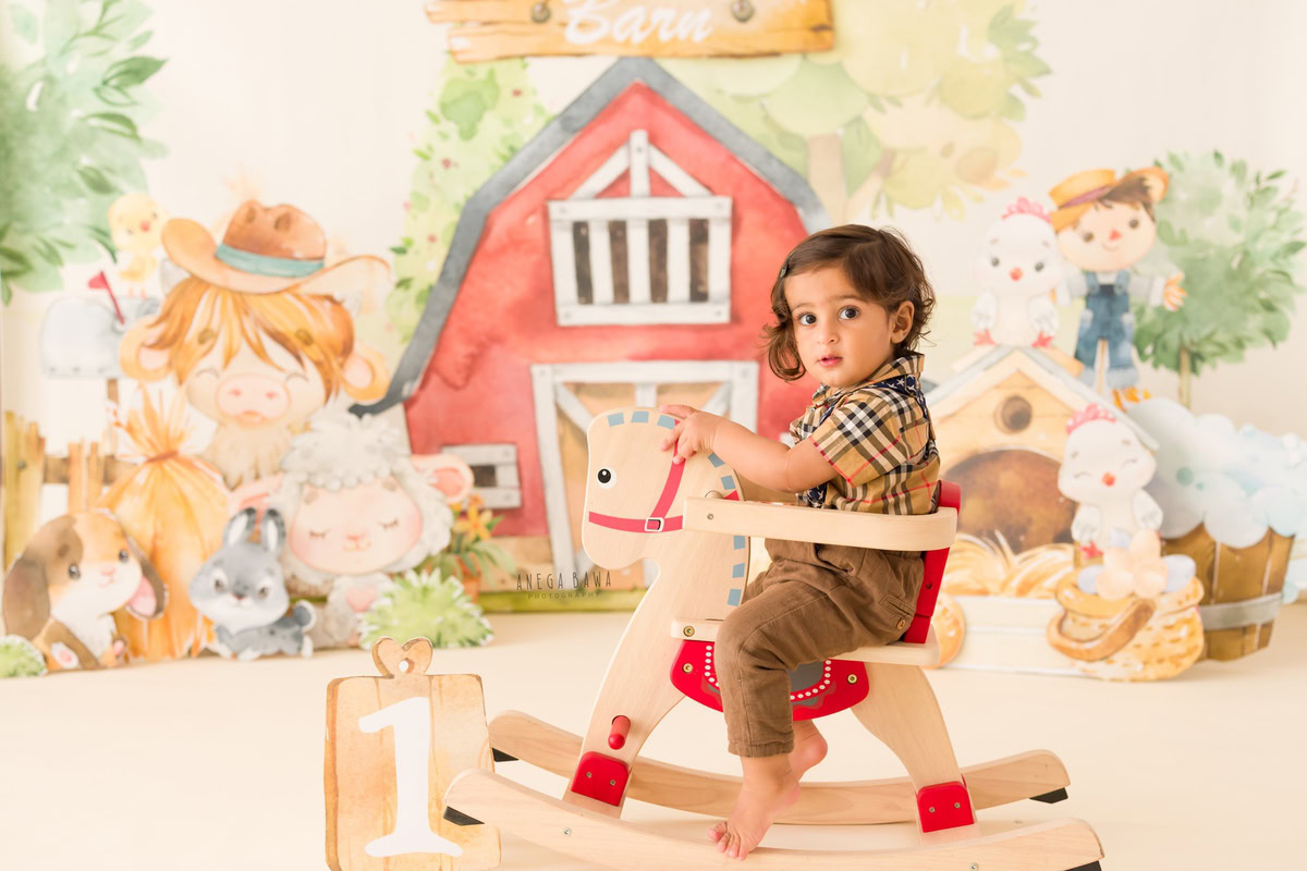 In this sitter birthday photoshoot by Anega Bawa in Delhi, the 9-month-old boy is captured sitting on a horse-shaped chair against a backdrop featuring playful cartoon motifs. The whimsical backdrop adds a fun and vibrant touch to the scene, creating a cheerful atmosphere for the birthday celebration. The boy's adorable pose and the colorful backdrop combine to make a delightful photograph, capturing the joy and excitement of this special milestone.