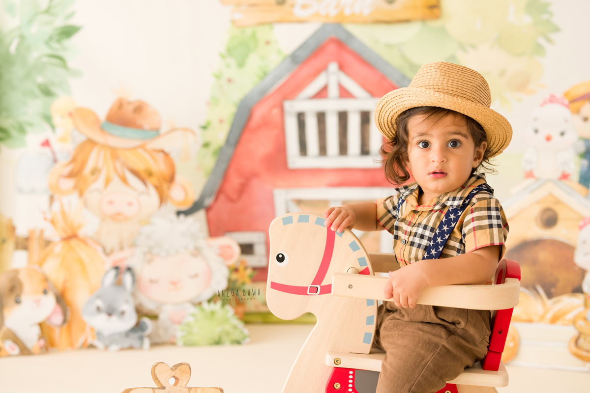 In this delightful sitter birthday photoshoot captured by Anega Bawa in Delhi, the 9-month-old boy is seated on a horse-shaped chair, adding a playful element to the scene. Adorned in a Burberry shirt and a hat, he exudes a charming and stylish vibe. The backdrop features colorful cartoon motifs, enhancing the cheerful and celebratory atmosphere of the occasion. The combination of the boy's adorable outfit, the whimsical backdrop, and his cheerful expression makes for a captivating photograph that beautifully commemorates this special milestone.