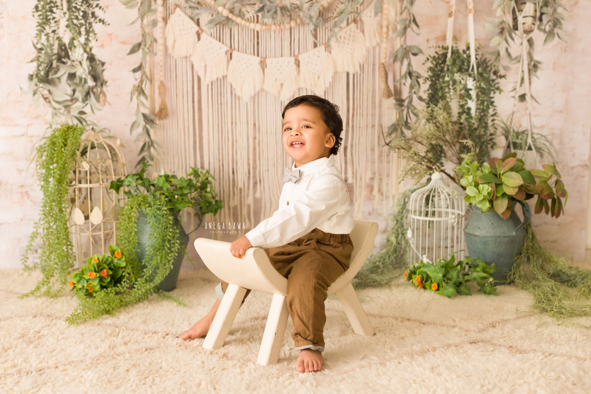At 9 months old, this little boy poses effortlessly on a slant chair against a chic beige backdrop, accented by delicate white castles and a vibrant green vase, all expertly captured by the talented sitter photographer Anega Bawa in Gurgaon.