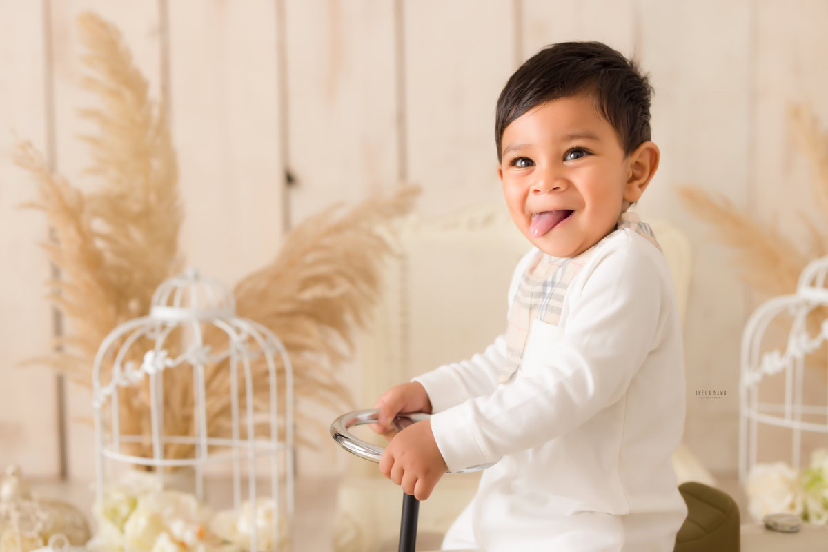 "9-months to 1-year-old boy gleefully sits on a toy scooter against a beige backdrop, surrounded by lush green bushes. Captured during a baby pre-birthday photoshoot by Anega Bawa, this charming moment radiates innocence and playful exploration."