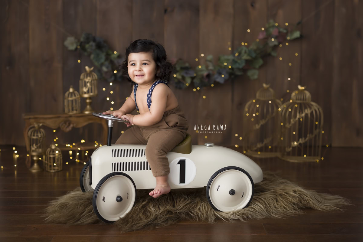 In a captivating photo captured by the skilled sitter photographer Anega Bawa in Gurgaon, this 9-month-old boy sits confidently on a toy scooter against a rich brown backdrop. The scene is illuminated by golden cages adorned with twinkling fairy lights, adding a magical touch to the image.