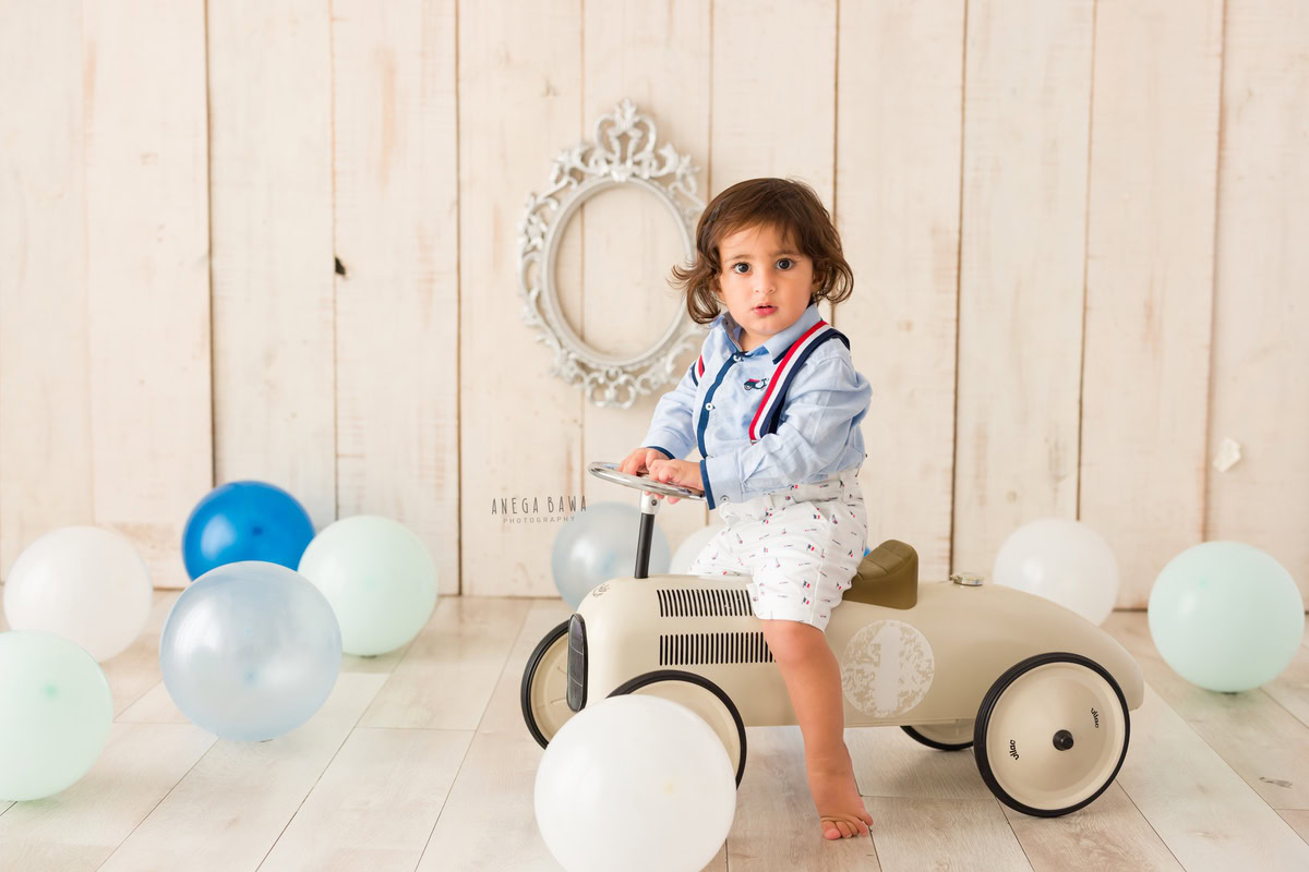 Delight in the innocence of your 9-month to 1-year-old with Anega Bawa's sitter photography in Delhi & Gurgaon. Your little one sits on a toy scooter amidst multicolour balloons, framed by a gleaming silver frame, against a soothing beige backdrop.