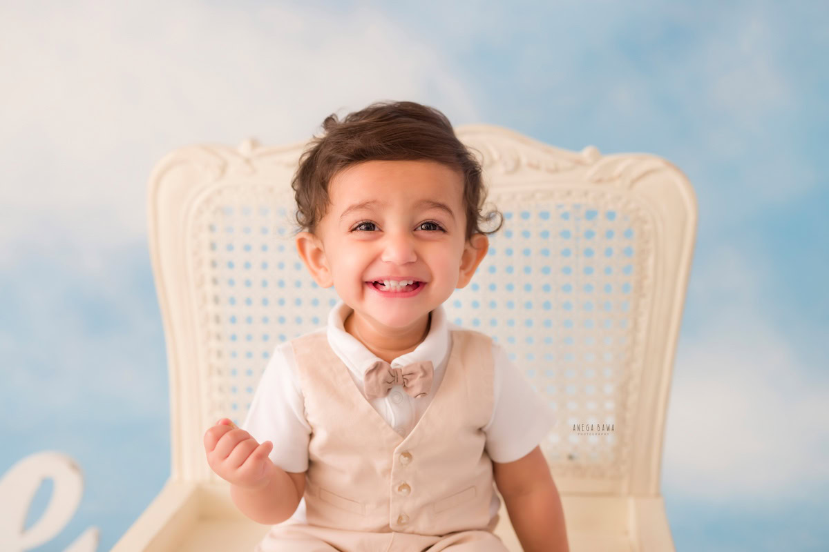 "Adorable 9-months to 1-year-old boy sitting on a white chair against a dreamy clouds backdrop. This heartwarming moment was captured during a baby pre-birthday photoshoot in Delhi, Gurgaon, and Noida by the talented Anega Bawa."
