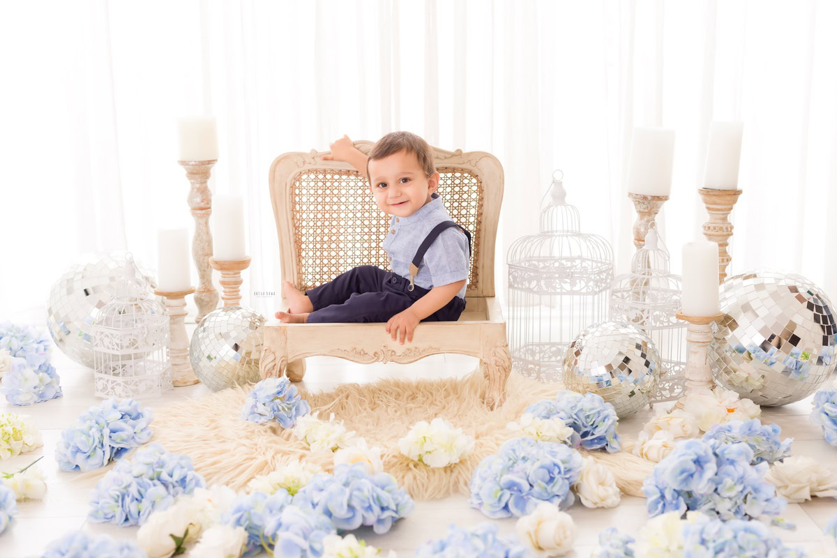 Boy sitting on a wooden chair surrounded by blue flowers, beside a candle stand and disco balls against a pristine white backdrop, marking his journey from 9 months to 1 year old during his first birthday photoshoot in Delhi by Anega Bawa, Gurgaon, Noida.