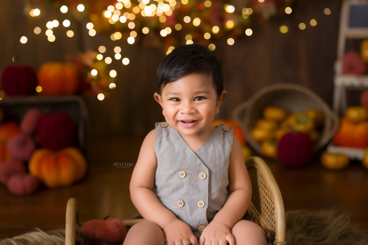 "Captivating image of a 9-months to 1-year-old boy sitting on a wooden cot, surrounded by enchanting fairy lights against a rustic brown backdrop. This delightful moment was captured during a baby pre-birthday photoshoot in Delhi, Gurgaon, and Noida by the skilled Anega Bawa."