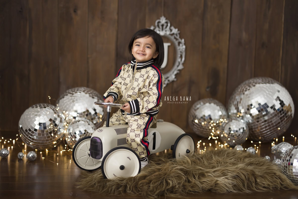 In this endearing toddler photoshoot orchestrated by Anega Bawa in Delhi, Gurgaon, and Noida, a boy, aged 9 months to 1 year old, sits on a toy scooter atop a brown rug. Behind him, a silver frame on the wall catches the eye amidst the enchanting glow of fairy lights against a brown backdrop. With each element thoughtfully curated, the scene captures the innocence and playful spirit of childhood, creating timeless memories for the family to treasure.