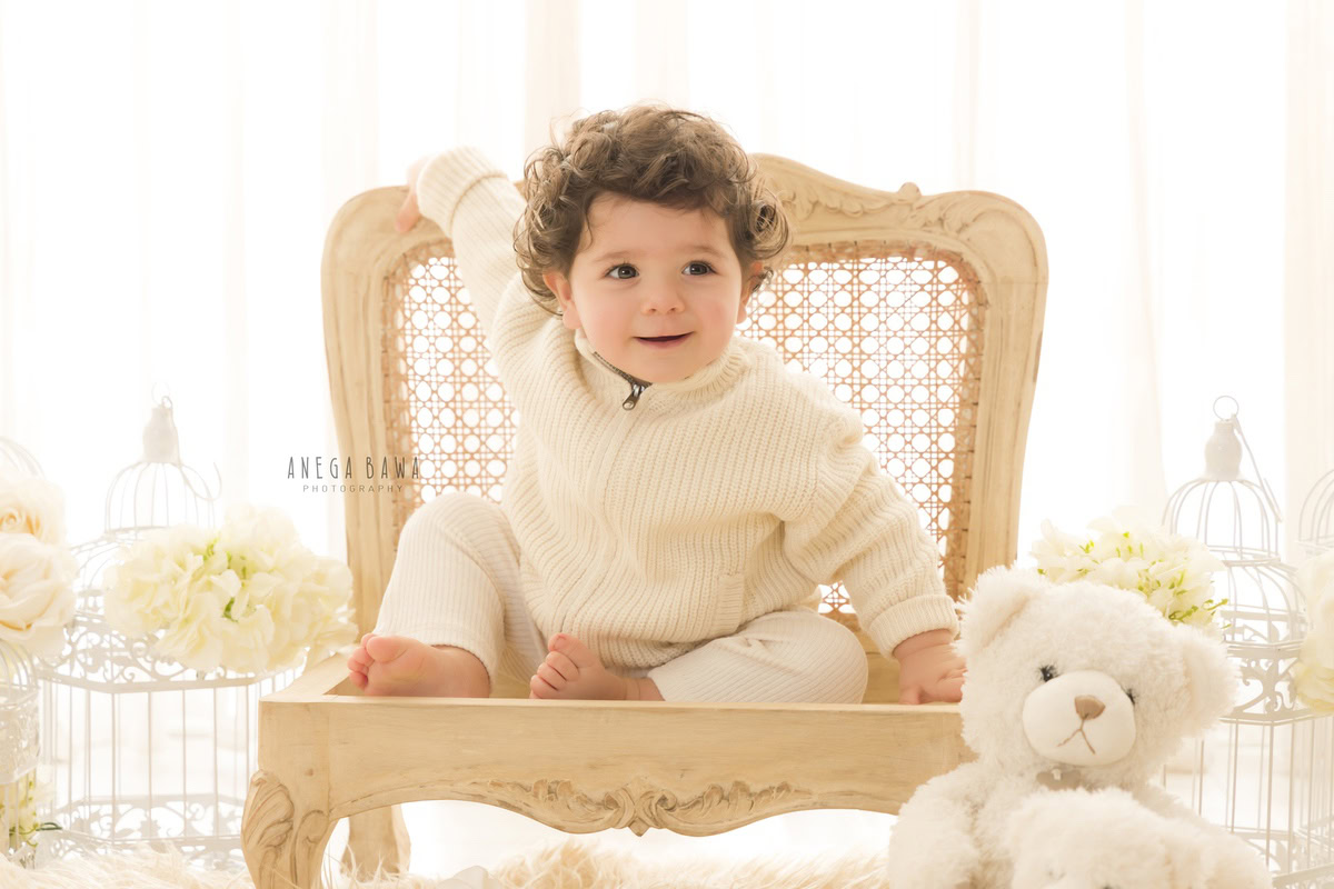 In this delightful toddler photoshoot by Anega Bawa in Delhi, Gurgaon, and Noida, a boy aged 10 months to 1 year old sits on a wooden chair surrounded by whimsical castles, white flowers, and cuddly teddy bears. Against a pristine white backdrop, each element adds to the enchanting ambiance, capturing the innocence and joy of childhood. These timeless moments are beautifully preserved, creating cherished memories for the family to treasure for years to come.
