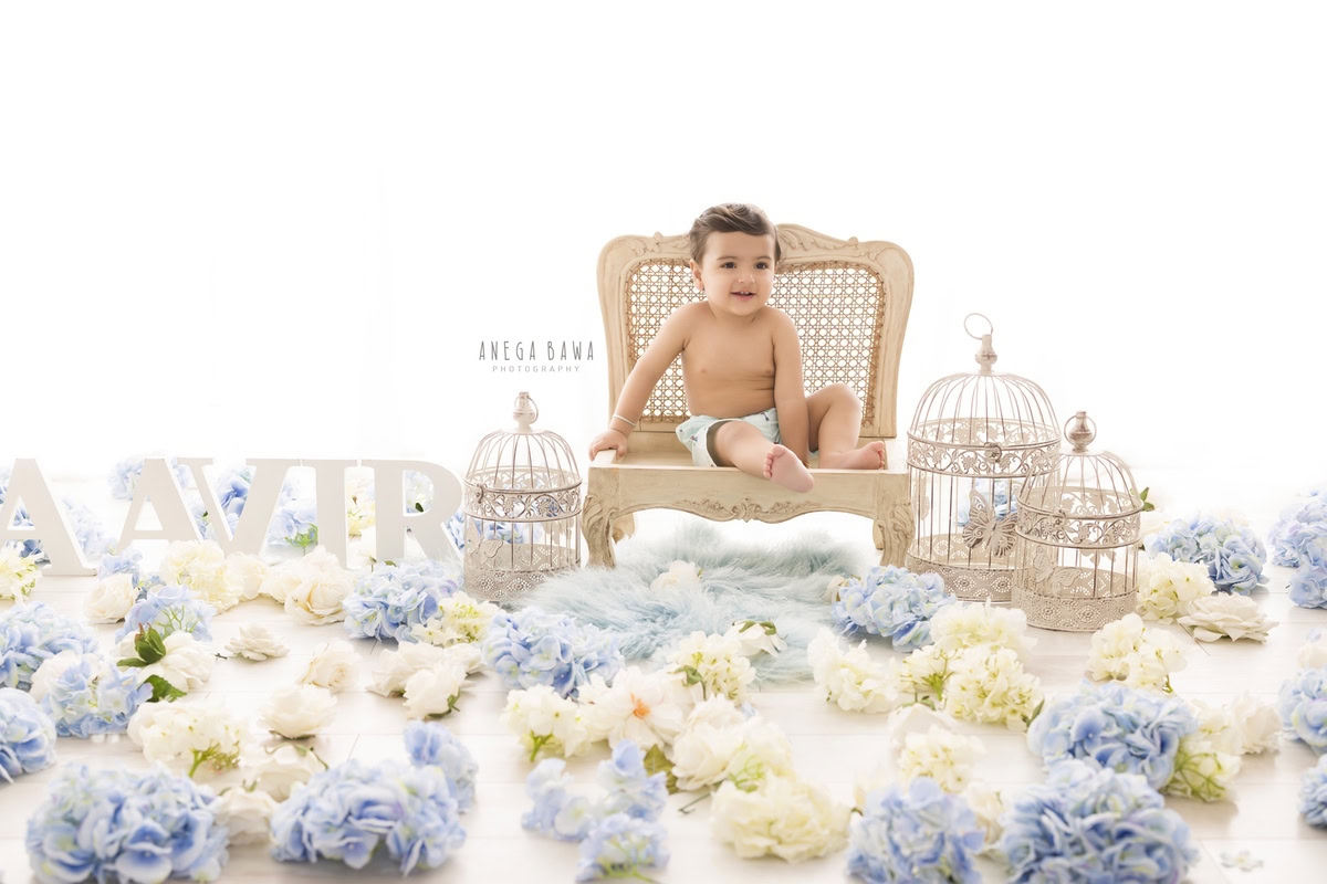In this delightful toddler photoshoot by Anega Bawa in Delhi, Gurgaon, and Noida, a 10-month to 1-year-old boy sits on a wooden chair amidst white castles and blue-white flowers. The serene white backdrop enhances the innocence, while a name frame on the floor adds a personal touch. These precious moments are expertly captured, preserving the beauty of childhood for the family to cherish forever.