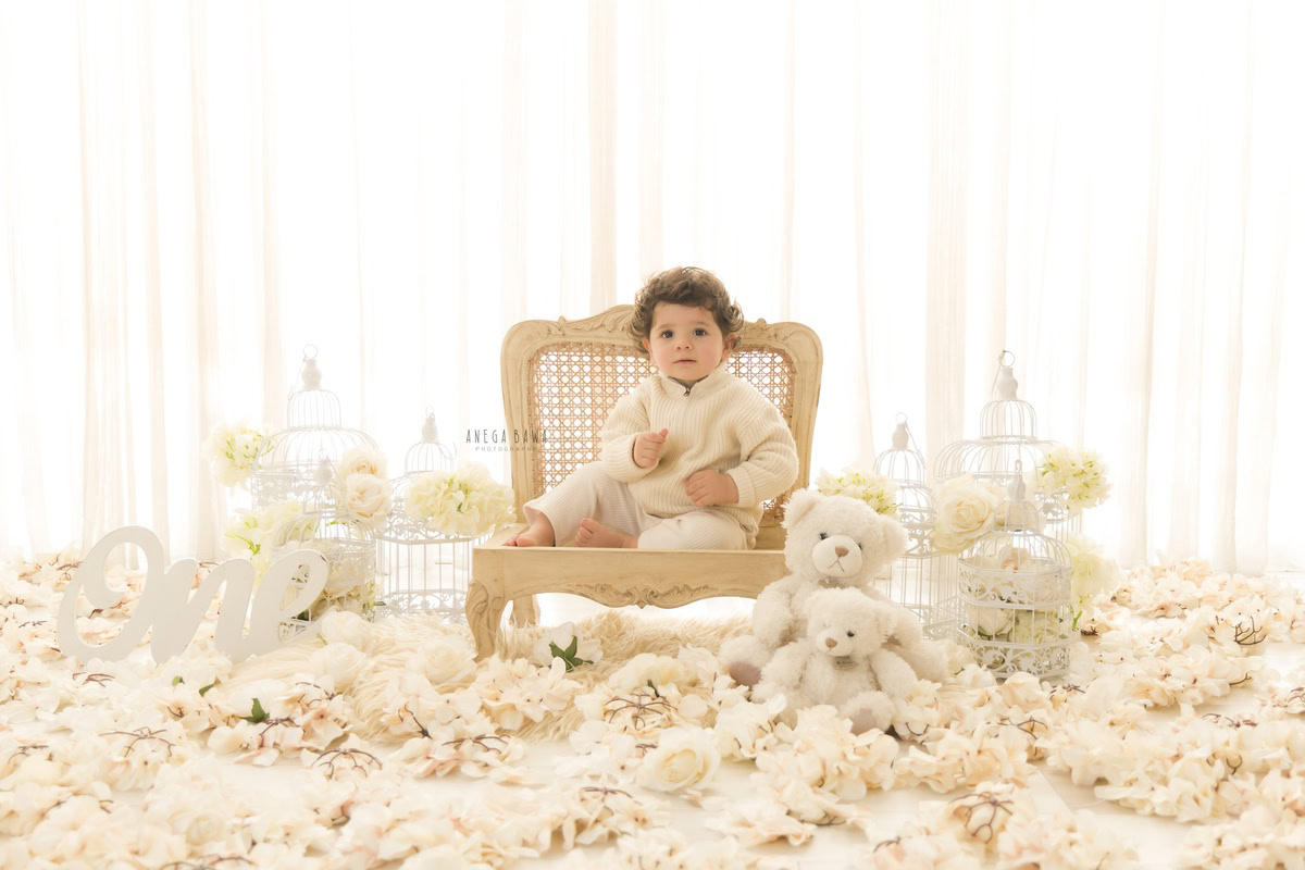 In this charming toddler photoshoot by Anega Bawa in Delhi, Gurgaon, and Noida, a 10-month to 1-year-old boy sits on a wooden chair surrounded by white flowers, castles, and teddy bears against a pristine white backdrop. These endearing moments are expertly captured, preserving the innocence and joy of childhood for the family to cherish forever.