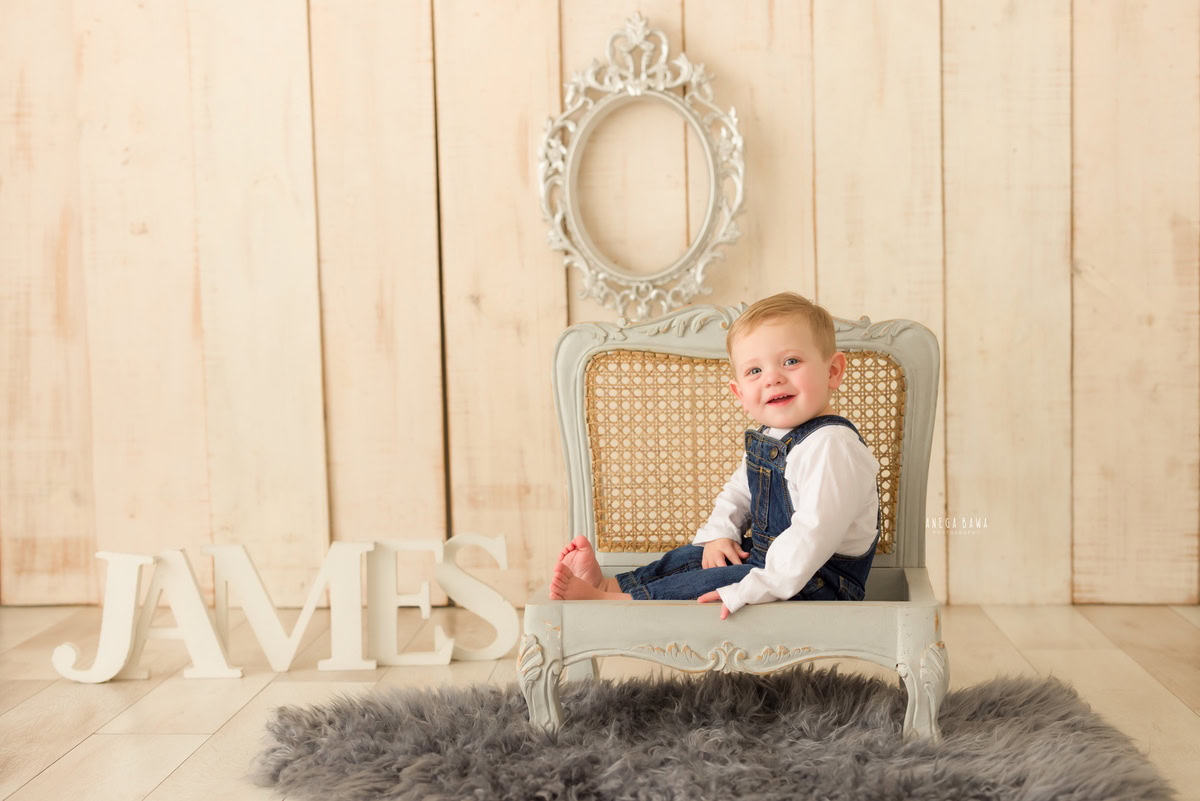 Charming 9-months to 1-year-old boy seated on a wooden grey chair, adorned with a grey rug and complemented by a grey frame on the wall. A name frame on the floor adds a personal touch to the scene, set against a warm beige backdrop. This captivating moment was captured during a baby pre-birthday photoshoot in Delhi, Anega Bawa, Gurgaon, and Noida.