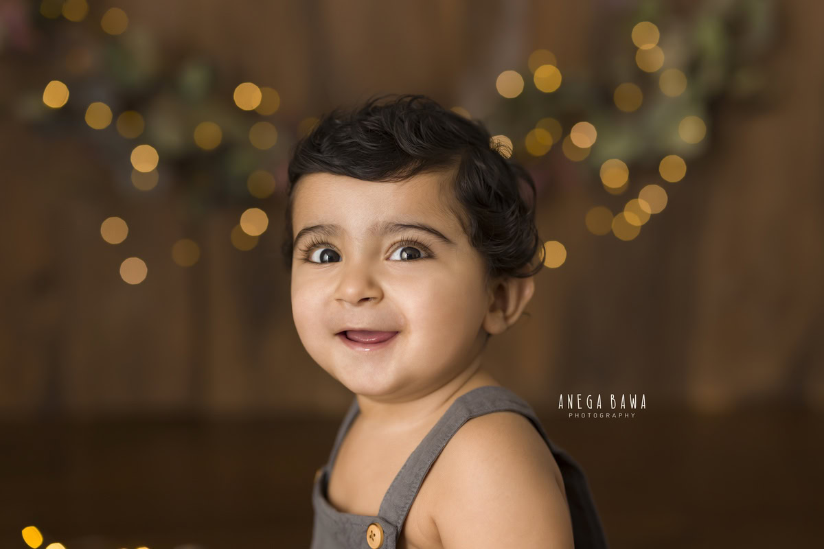 In this delightful toddler photoshoot by Anega Bawa in Delhi, Gurgaon, and Noida, a 9-month to 1-year-old boy beams with joy against a warm brown backdrop illuminated by golden lights. Each smile captured in this session is a precious memory, encapsulating the happiness and innocence of childhood for years to come.