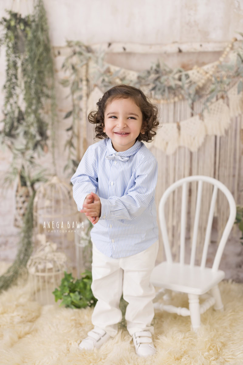 Boy standing on a cozy beige rug beside a white chair, with leafy fringes on the wall, commemorating his journey from 8 months to 1 year old during his first birthday photoshoot in Delhi by Anega Bawa, Gurgaon, Noida.