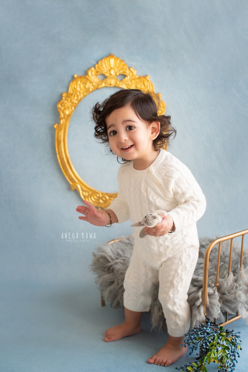Celebrate the enchanting moments of your little boy's journey with Anega Bawa Photography in Delhi and Gurgaon. Adorned in a golden cot against a serene blue backdrop, complemented by a golden frame on the wall, your 8-month to 1-year-old stands in captivating elegance, creating timeless memories to cherish forever.