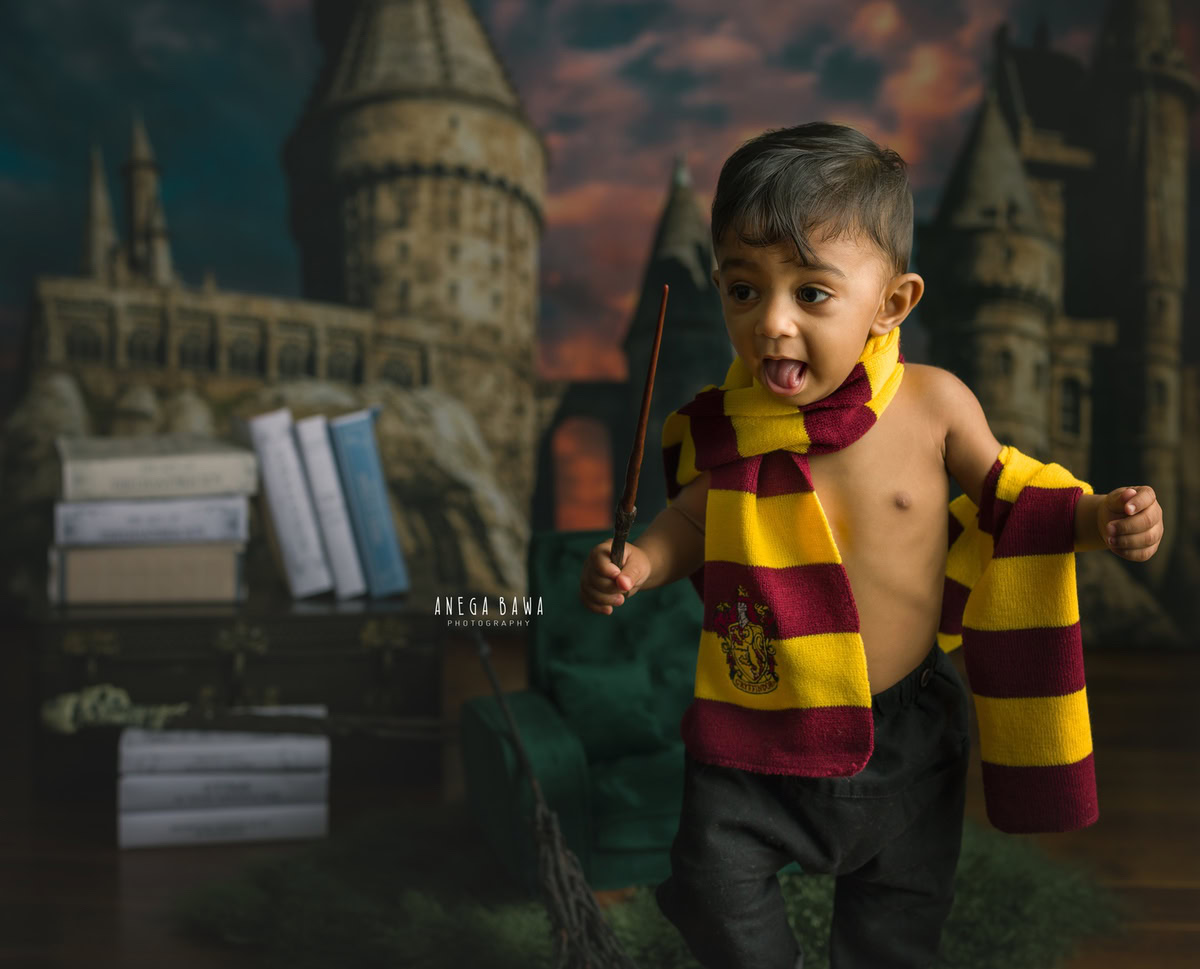 Capture the magic of childhood with Anega Bawa's sitter photography in Delhi & Gurgaon. Your 8-month to 1-year-old stands tall, adorned in a Harry Potter-inspired look, amidst a whimsical bookshelf backdrop under the warm glow of orange clouds.