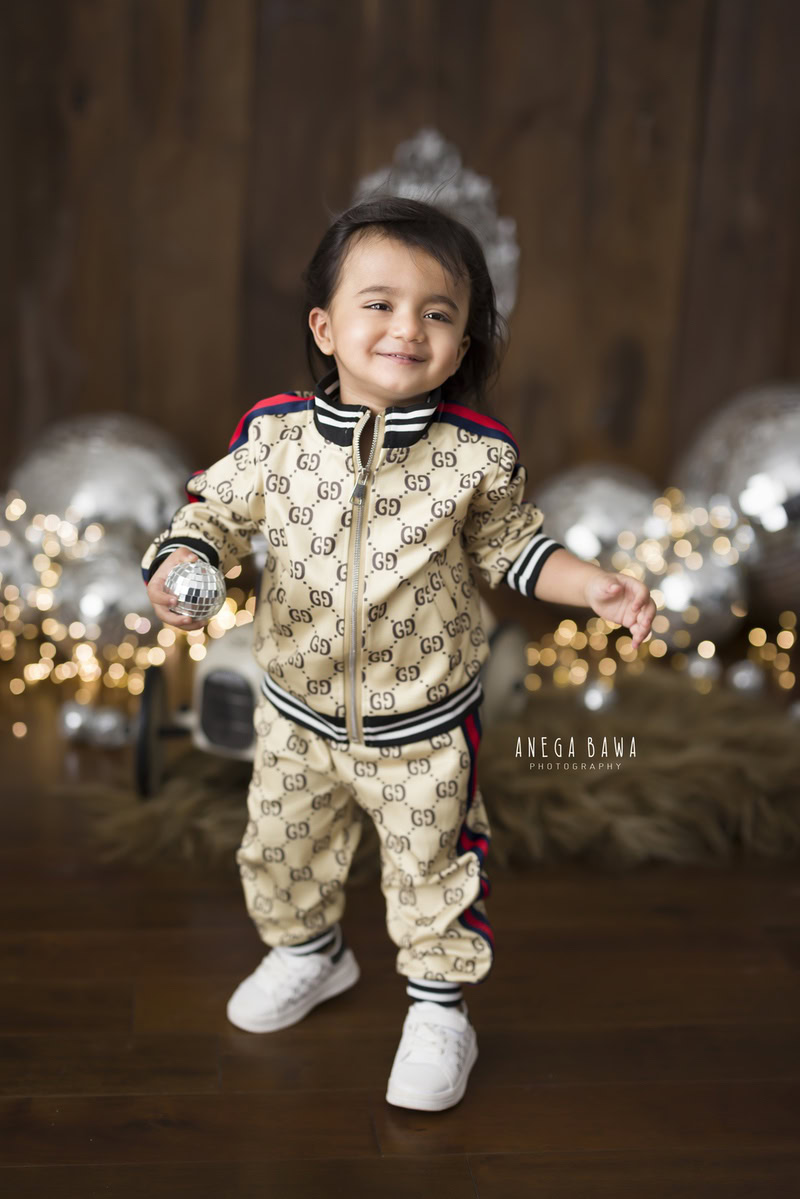In this enchanting toddler photoshoot captured by Anega Bawa in Delhi, Gurgaon, and Noida, a charming 8-month to 1-year-old boy stands tall, holding disco balls against a rich brown backdrop. Each moment frozen in time during this session preserves the curious spirit and boundless energy of childhood, creating memories to cherish for a lifetime.