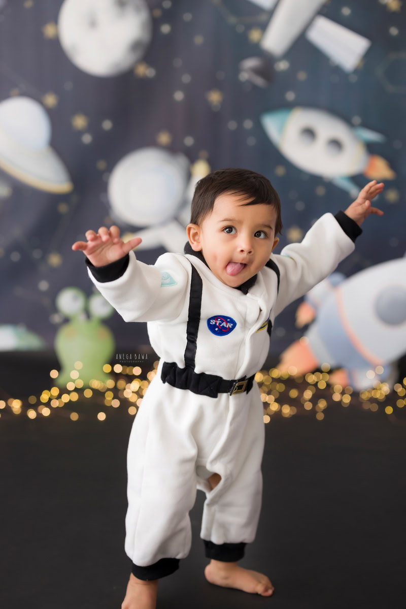 8-months to 1-year-old boy standing in a charming space outfit, set against a space-themed backdrop illuminated by fairy lights. This captivating moment was captured during a baby pre-birthday photoshoot in Delhi, Anega Bawa, Gurgaon, and Noida.