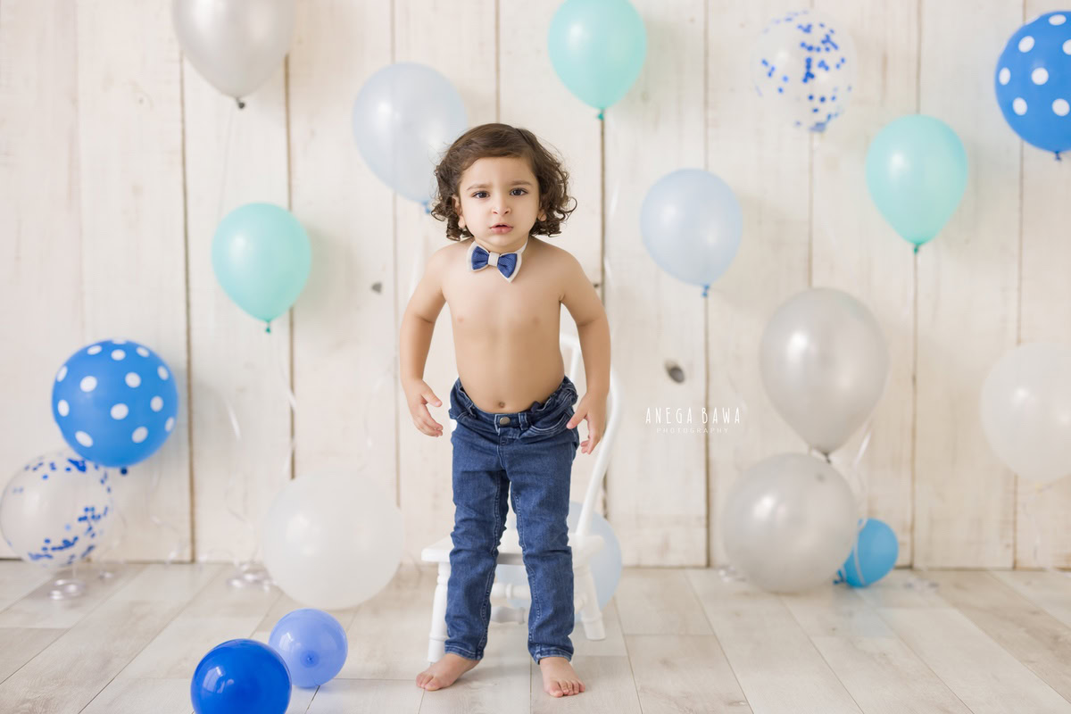 Boy standing amidst multicolor balloons wearing a cute bow and denim outfit against a serene beige backdrop, capturing his journey from 8 months to 1 year old during his first birthday photoshoot in Delhi by Anega Bawa, Gurgaon, Noida.