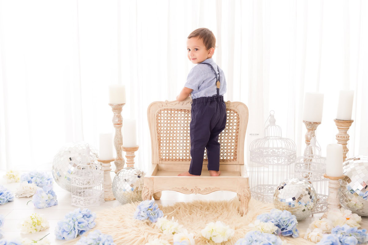 Boy standing on a wooden chair surrounded by candle stands, disco balls, blue and white flowers against a pristine white backdrop, marking his journey from 8 months to 1 year old during his first birthday photoshoot in Delhi by Anega Bawa, Gurgaon, Noida.