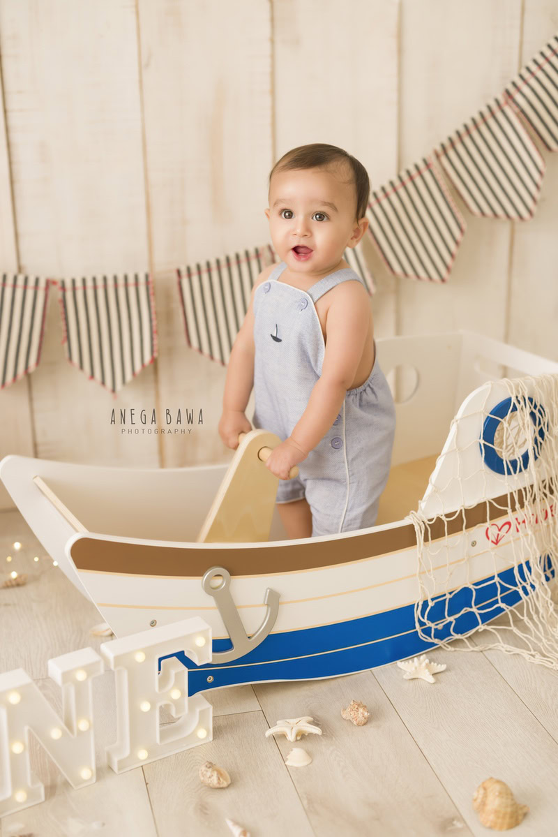 Boy standing beside a toy spaceship against a serene beige backdrop with petals scattered on the floor, marking his journey from 8 months to 1 year old during his first birthday photoshoot in Delhi by Anega Bawa, Gurgaon, Noida.
