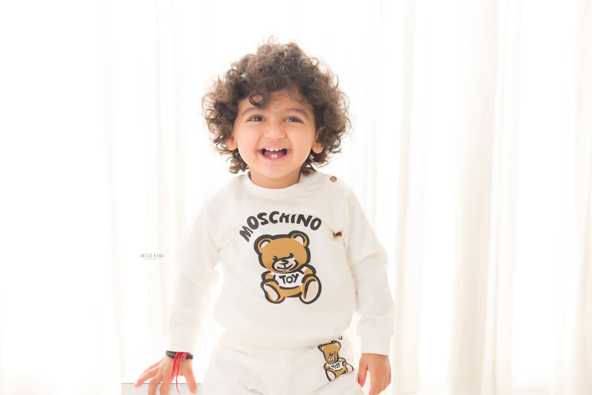 Immerse in the charm of your little one with Anega Bawa's sitter photography in Delhi & Gurgaon. Your 8-month to 1-year-old stands proudly against a clean white backdrop, showcasing adorable curly hair, capturing timeless moments.