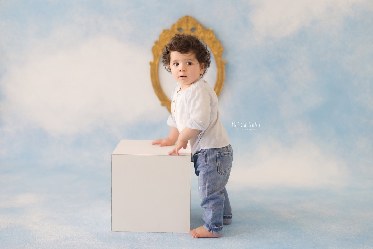 Witness the grace of your little one with Anega Bawa's sitter photography in Delhi & Gurgaon. Standing elegantly against a cloudy backdrop, your 8-month to 1-year-old shines beside a golden frame on the wall, creating captivating memories.