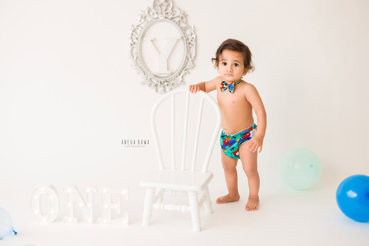 In this delightful photo, an 8-months to 1-year-old boy stands beside a white chair, dressed in cute bow shorts. The backdrop features an alphabet frame against a pristine white backdrop, creating a charming scene. This adorable moment was captured during a baby pre-birthday photoshoot in Delhi, Anega Bawa, Gurgaon, and Noida.