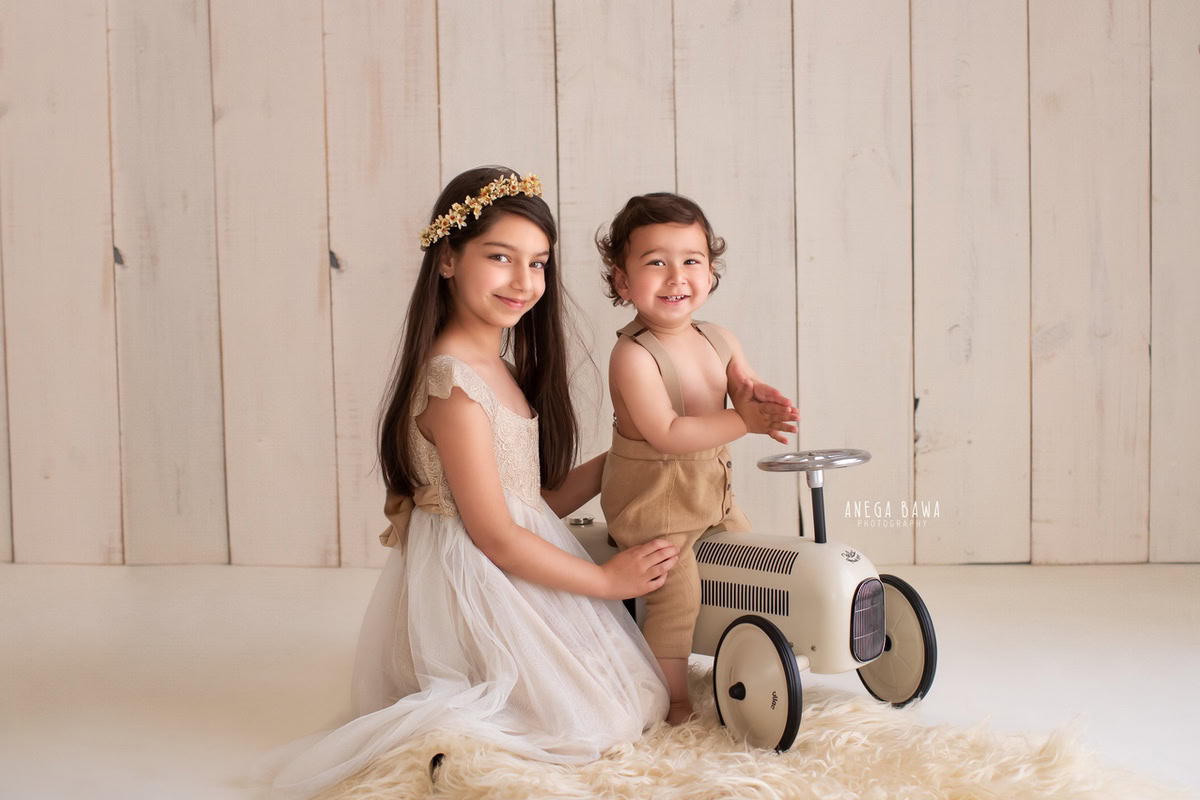 In an enchanting portrait by the talented sitter photographer Anega Bawa in Gurgaon, a lively 8-month-old boy is captured alongside his sibling, both gleefully playing with a toy scooter. Against a soothing beige backdrop and rug, their joyous camaraderie shines through, creating a heartwarming moment frozen in time.
