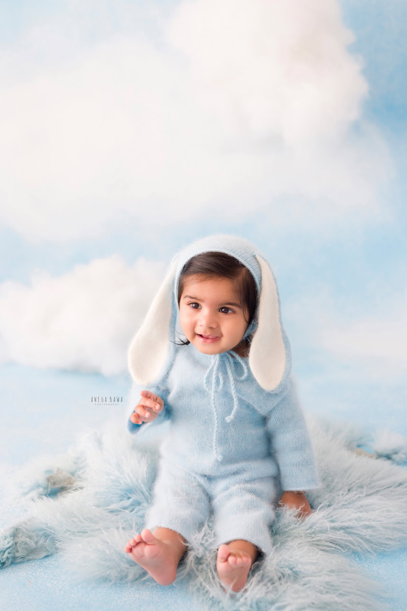 In this charming sitter birthday photoshoot, the 8-month-old boy wears a blue babysuit paired with a cute cap. Seated on a blue rug against a cloudy backdrop, he exudes innocence and joy. The soft atmosphere adds whimsy to the scene, highlighting his playful demeanor and curious gaze. This delightful photograph captures the essence of childhood wonder and marks a special milestone in his journey.