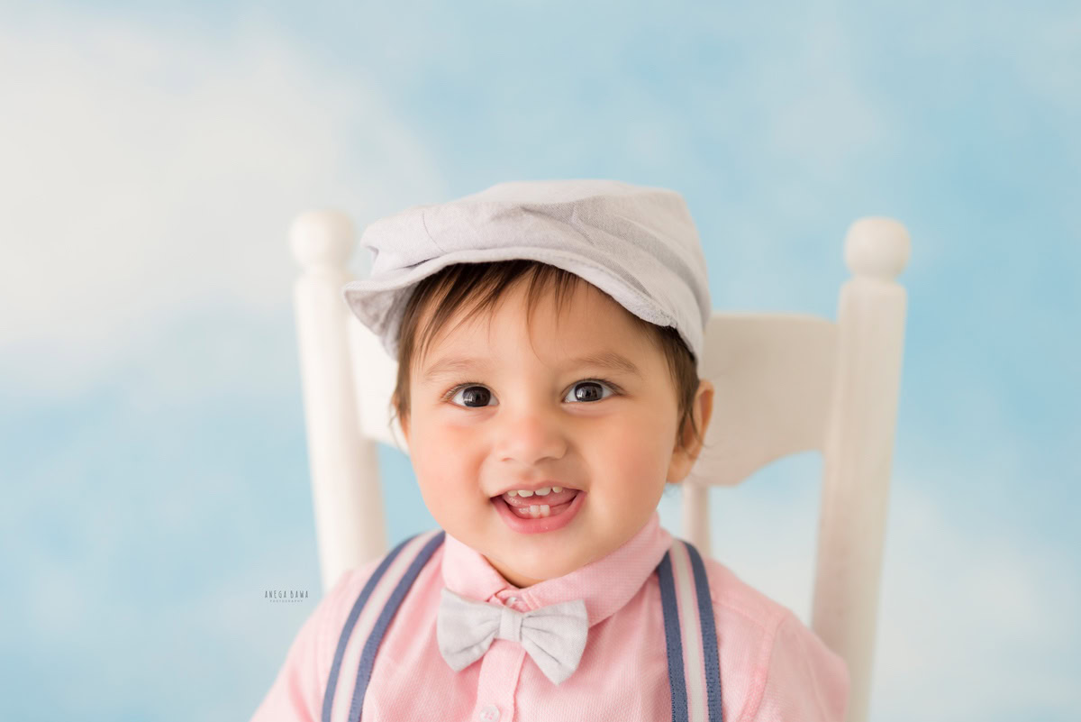 Capture the adorable essence of your little one with Anega Bawa's sitter photography in Delhi & Gurgaon. Dressed in a charming bow cap, he sits gracefully on a white chair against a serene cloud backdrop. Perfect for 8-month to 1-year-old sitters, this session promises enchanting memories to cherish forever.