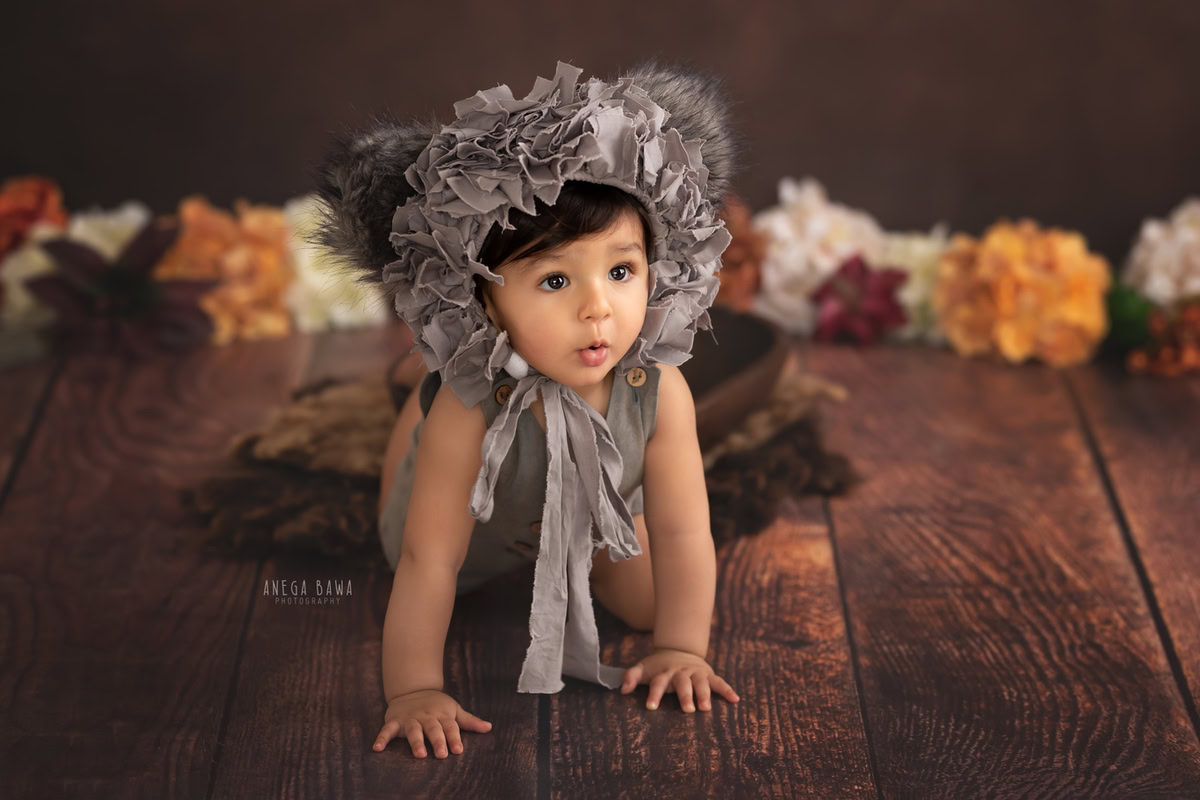 In this captivating photograph captured by the skilled sitter photographer Anega Bawa in Gurgaon, a charming 12-month-old boy is adorned with a stylish grey unique cap, adding a touch of flair to his ensemble. Set against a rich brown backdrop adorned with delicate flowers, the image exudes a timeless charm and captures the innocence and wonder of childhood.