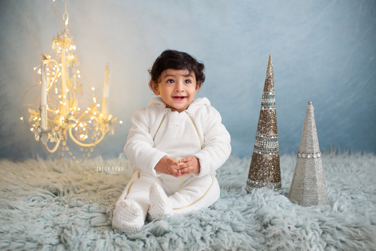 In this captivating sitter photography session by Anega Bawa in Delhi, Gurgaon, a charming 12-month-old boy dons a pristine white babysuit as he sits on a cozy blue rug. The scene is illuminated by the warm glow of candlestands and golden lights against a serene blue backdrop, creating a magical ambiance. This adorable moment captures the innocence and wonder of childhood, making it a cherished memory for years to come.