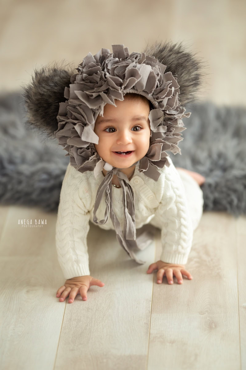 Capture the innocence and charm of your little one with Anega Bawa Photography in Delhi and Gurgaon. Adorned in a white babysuit and a unique grey cap, seated on a soft grey rug against a pristine white backdrop, your 12-month to 1-year-old shines in every frame, creating timeless memories.