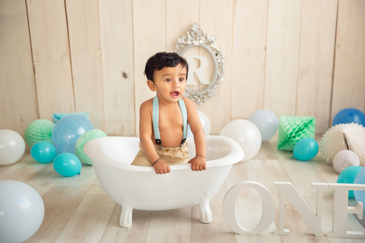 Capture the adorable charm of your little one in a delightful photoshoot by Anega Bawa Photography in Delhi and Gurgaon. Adorned with twinkling fairy lights and surrounded by vibrant blue and green pompom balloons, your 12-month to 1-year-old prince sits in a pristine white tub against a soothing beige backdrop, with a shimmering silver alphabet frame adding a touch of elegance to the scene.