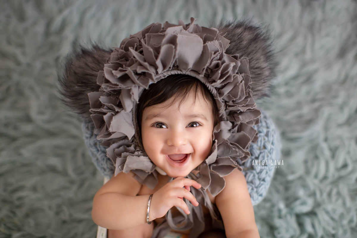 Captured in this delightful sitter photoshoot by Anega Bawa in Delhi, Gurgaon, is a joyful 12-month-old boy sporting a unique cap. Seated on a soft grey rug, his infectious smile lights up the scene, creating a heartwarming moment frozen in time. This charming pose perfectly encapsulates the innocence and happiness of childhood, making it a treasured memory for years to come.