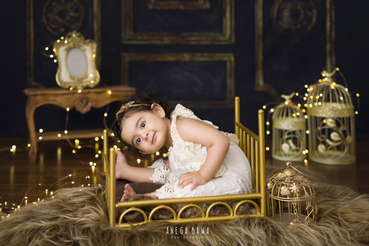In this enchanting photograph by Anega Bawa, a 15-month-old girl sits on a cute golden cot, surrounded by golden castles and delicate frames, creating a magical ambiance. The warm hues of the brown backdrop complement the scene beautifully, while fairy lights add a touch of whimsy. Anega Bawa's skilled photography captures the innocence and charm of the girl at 15 months, creating a captivating image that will be cherished for years to come.