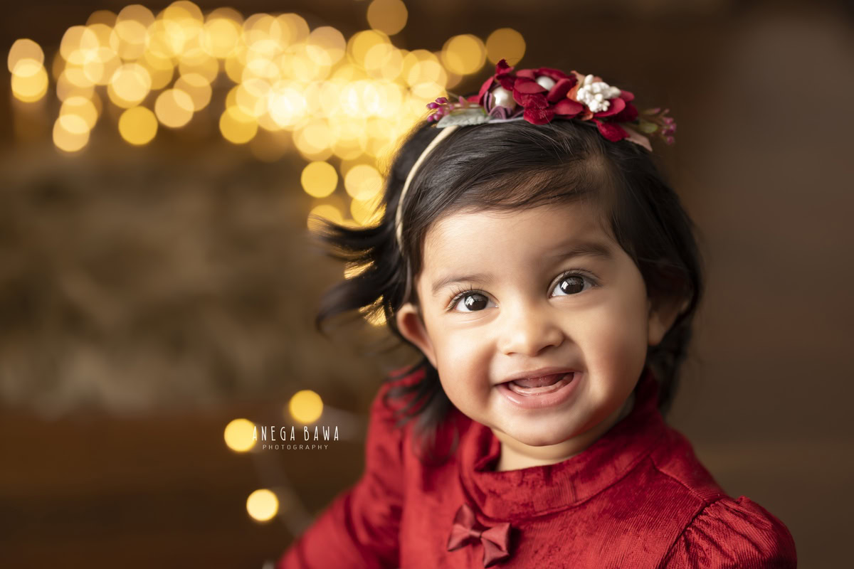 In this delightful toddler photoshoot by Anega Bawa in Delhi, Gurgaon, and Noida, a precious 12-month-old girl sits gracefully against a warm brown backdrop adorned with golden lights. She wears a radiant red dress and a matching headband, adding a pop of color and charm to the scene. Each snapshot captures the innocence and beauty of childhood, creating timeless memories for the family to cherish.