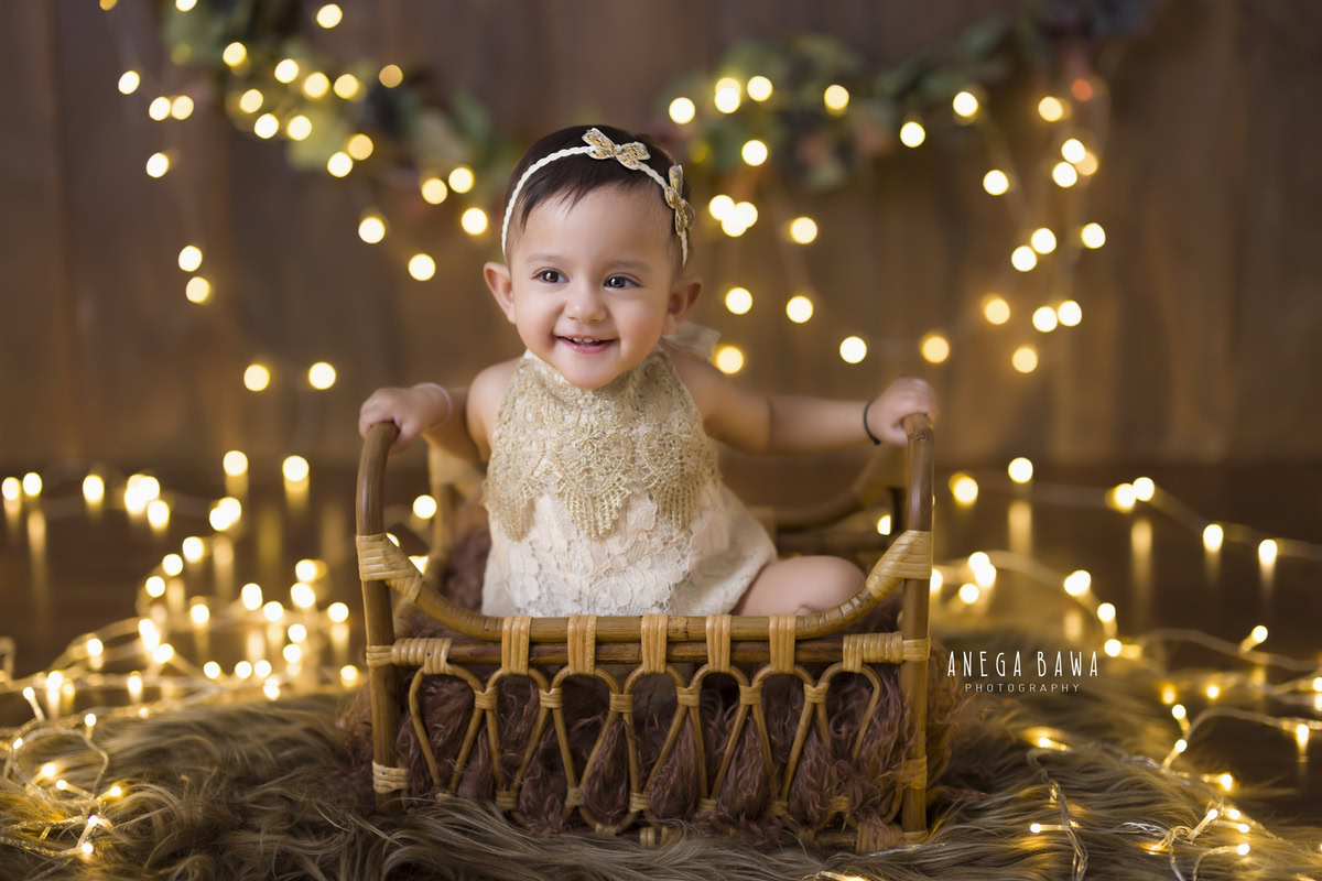In this enchanting toddler photoshoot by Anega Bawa in Delhi, Gurgaon, and Noida, a lovely 12-month-old girl sits elegantly on a wooden brown cot against a warm brown backdrop illuminated by golden lights. She wears a stunning golden dress, complementing the rich hues of the setting. The brown rug adds a cozy touch to the scene, creating a serene and captivating atmosphere. Each photograph captures the essence of childhood innocence and beauty, ensuring cherished memories for years to come.