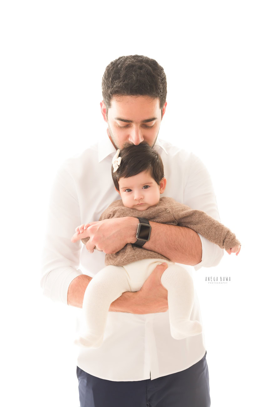 Heartwarming family photoshoot by Anega Bawa Photography, the renowned photographer in Delhi and Gurgaon, featuring a precious 3, 4, 5, 6-month-old baby girl being kissed by her father against a pure white backdrop.