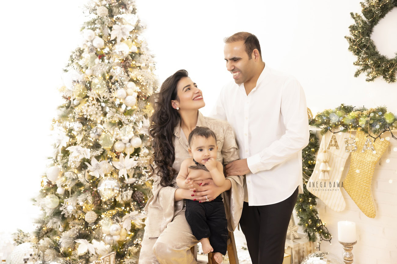 Charming family photoshoot by Anega Bawa Photography, the renowned photographer in Delhi and Gurgaon, featuring a delightful 3, 4, 5, 6-month-old baby with Mom and Dad, a Christmas tree, and Santa boots against a pure white backdrop.
