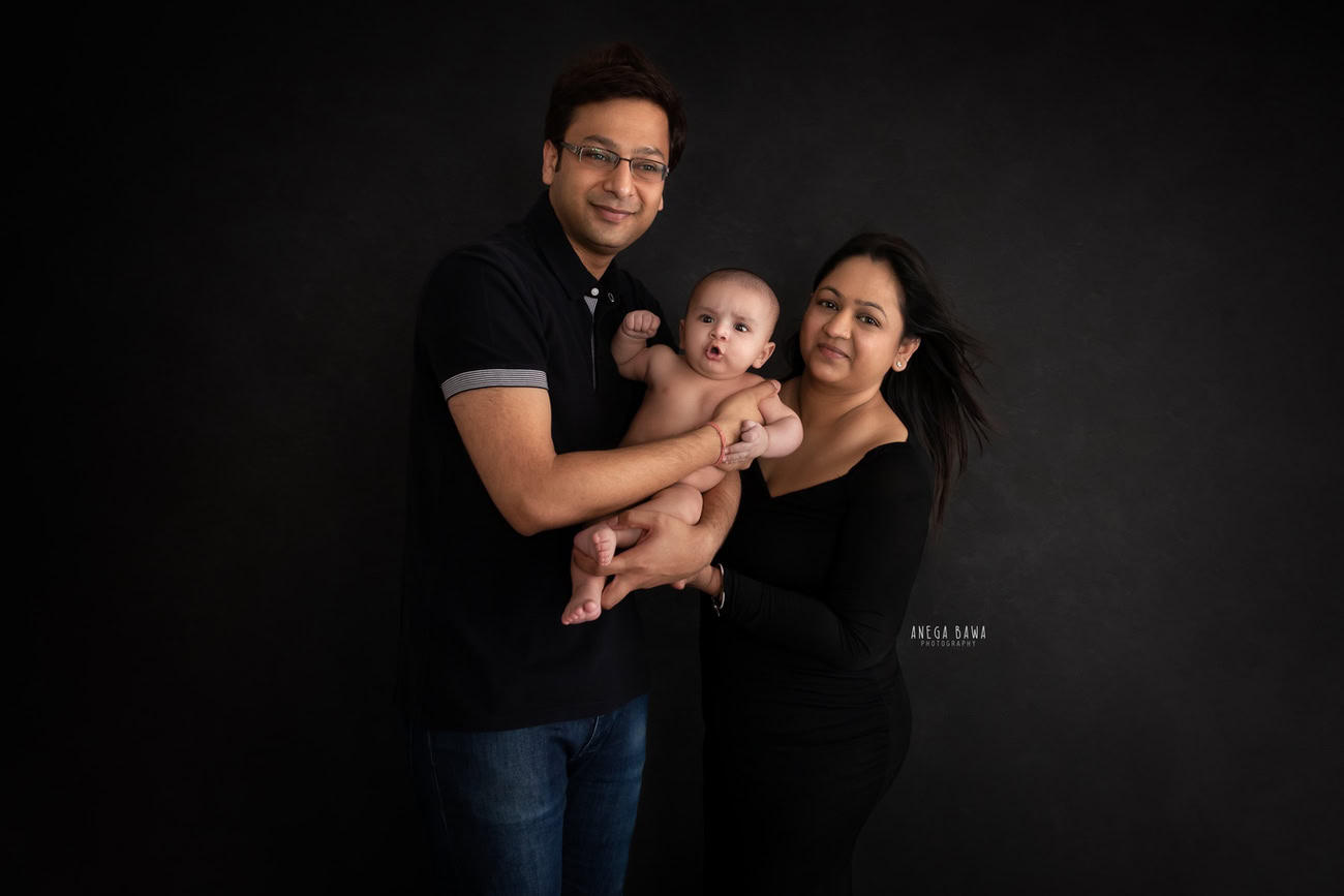 Elegant family photoshoot by Anega Bawa Photography, the renowned photographer in Delhi and Gurgaon, featuring a precious 3, 4, 5, 6-month-old baby boy with Mom and Dad against a sophisticated black backdrop.
