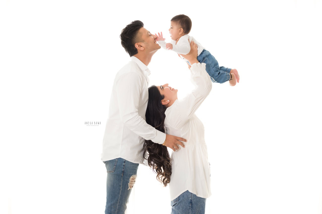 Elegant family photoshoot by Anega Bawa Photography, the renowned photographer in Delhi and Gurgaon, featuring a precious 3, 4, 5, 6-month-old baby daughter with Mom and Dad, all dressed in white shirts, against a pure white backdrop.