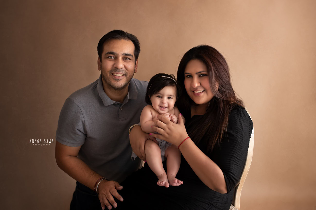Captivating family photoshoot by Anega Bawa Photography, the esteemed photographer in Delhi and Gurgaon, showcasing the endearing bond between Mom, Dad, and their precious 3, 4, 5, 6-month-old daughter against a warm dusky brown backdrop.