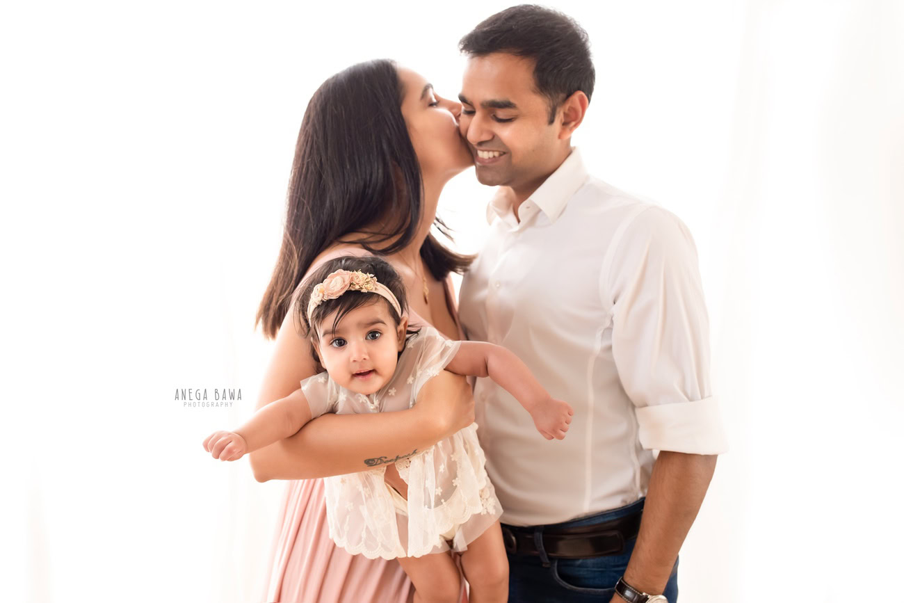 Cherished family photoshoot captured by Anega Bawa Photography, the esteemed photographer in Delhi and Gurgaon, featuring the loving bond between Mom, Dad, and their precious 3, 4, 5, 6-month-old daughter in a tender kissing pose against a serene white backdrop.