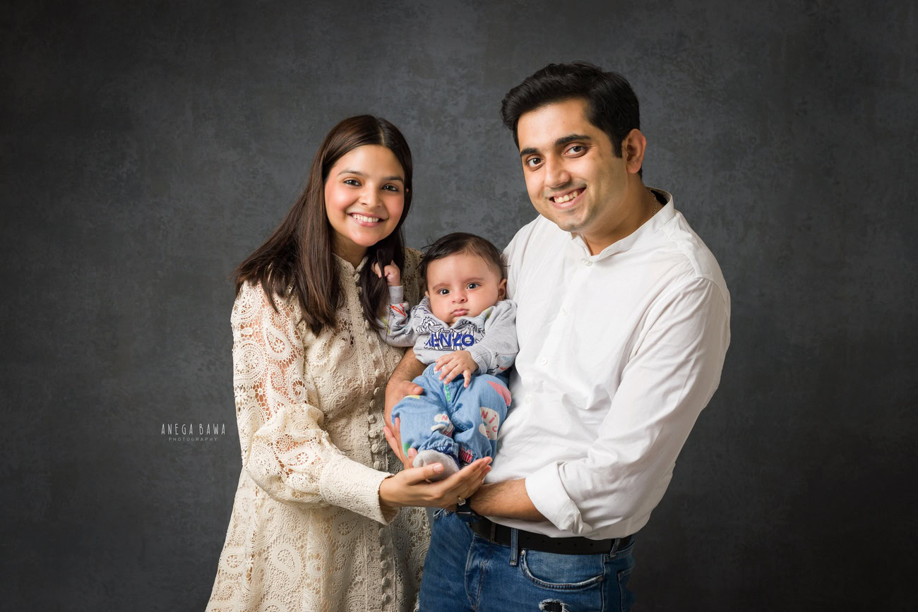 Captivating family photoshoot by Anega Bawa Photography, the esteemed photographer in Delhi and Gurgaon, portraying the endearing bond of Mom and Dad holding their beloved 3, 4, 5, 6-month-old son against a stylish grey backdrop.
