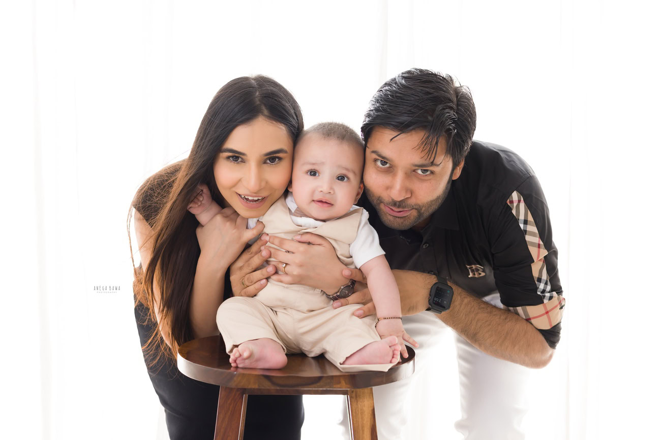 Captivating family photoshoot by Anega Bawa Photography, the esteemed photographer in Delhi and Gurgaon, capturing the endearing moments of Mom, Dad, and their precious 3, 4, 5, 6-month-old son sitting on a wooden stool against a serene white backdrop.