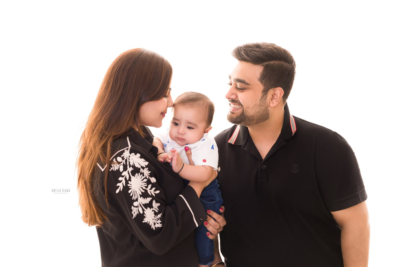 Captivating family photoshoot by Anega Bawa Photography, the esteemed photographer in Delhi and Gurgaon, capturing the joyous moments of Mom, Dad, and their adorable 3, 4, 5, 6-month-old son in a smiling pose against a serene white backdrop.